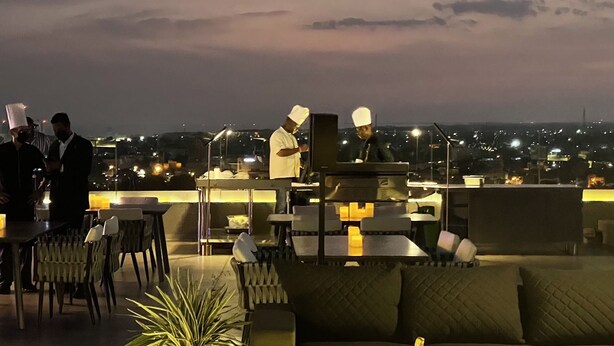 A rooftop lounge with outdoor seating, a chef preparing food, and a cityscape view at dusk. - CIKA Golden Hotel and Suites (Lomé)