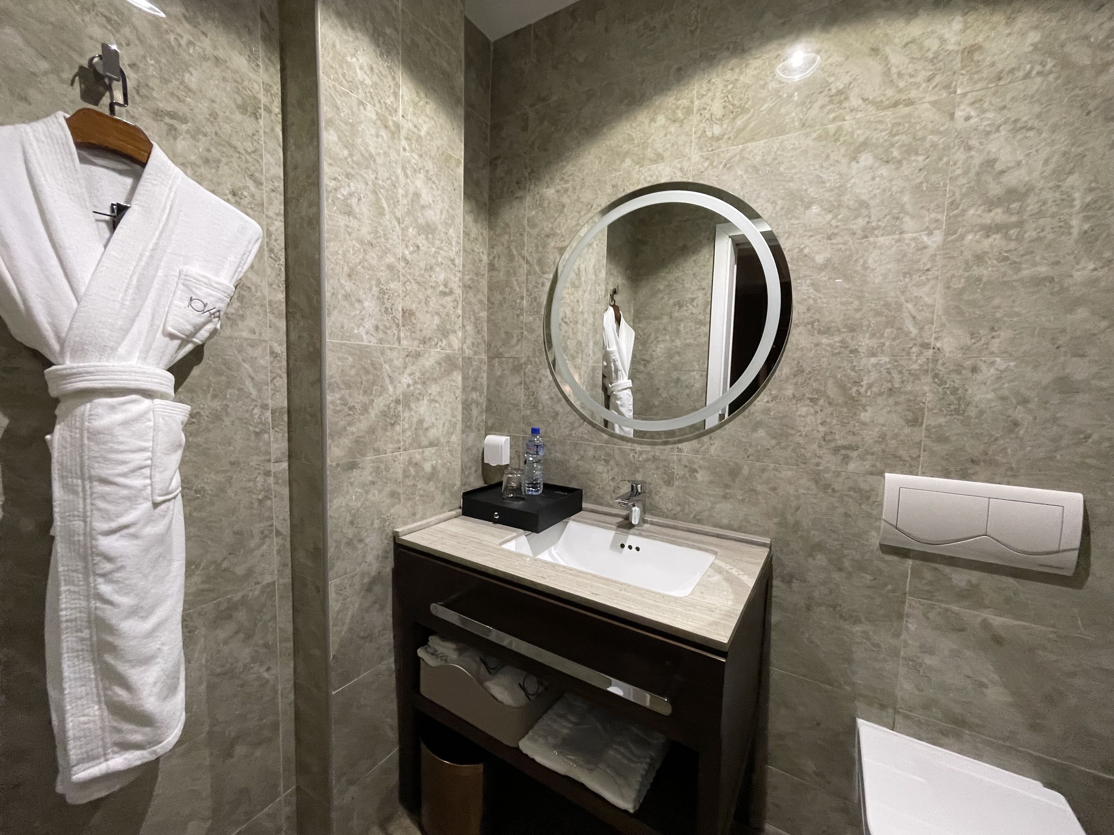 A bathroom with a marble wall, a round mirror, a sink, and a robe hanging on a rack.
