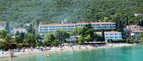 A coastal hotel with multiple floors, a beach in the foreground, and a forested hill in the background.