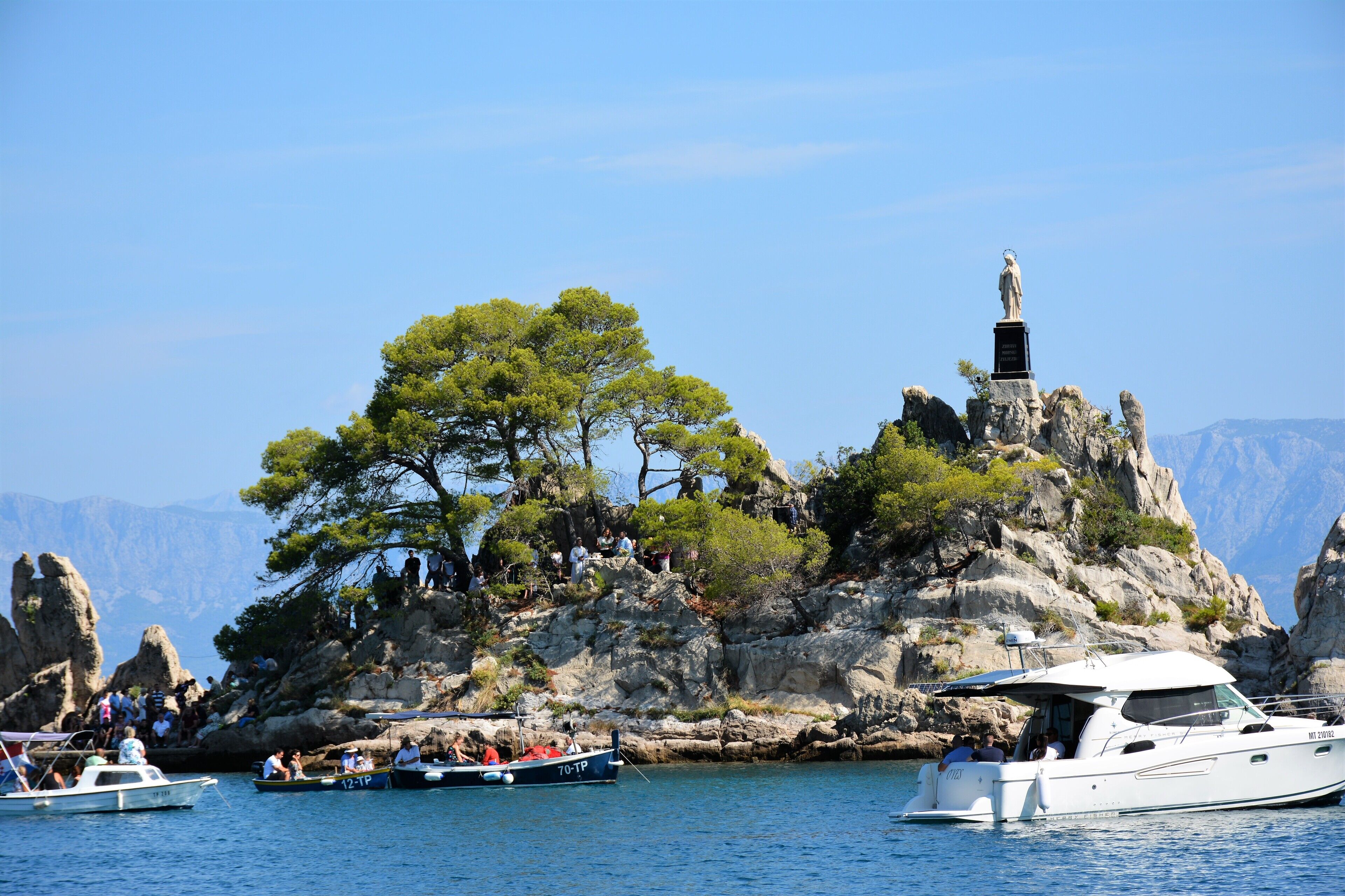 A rocky island with a statue, surrounded by clear blue water and boats.