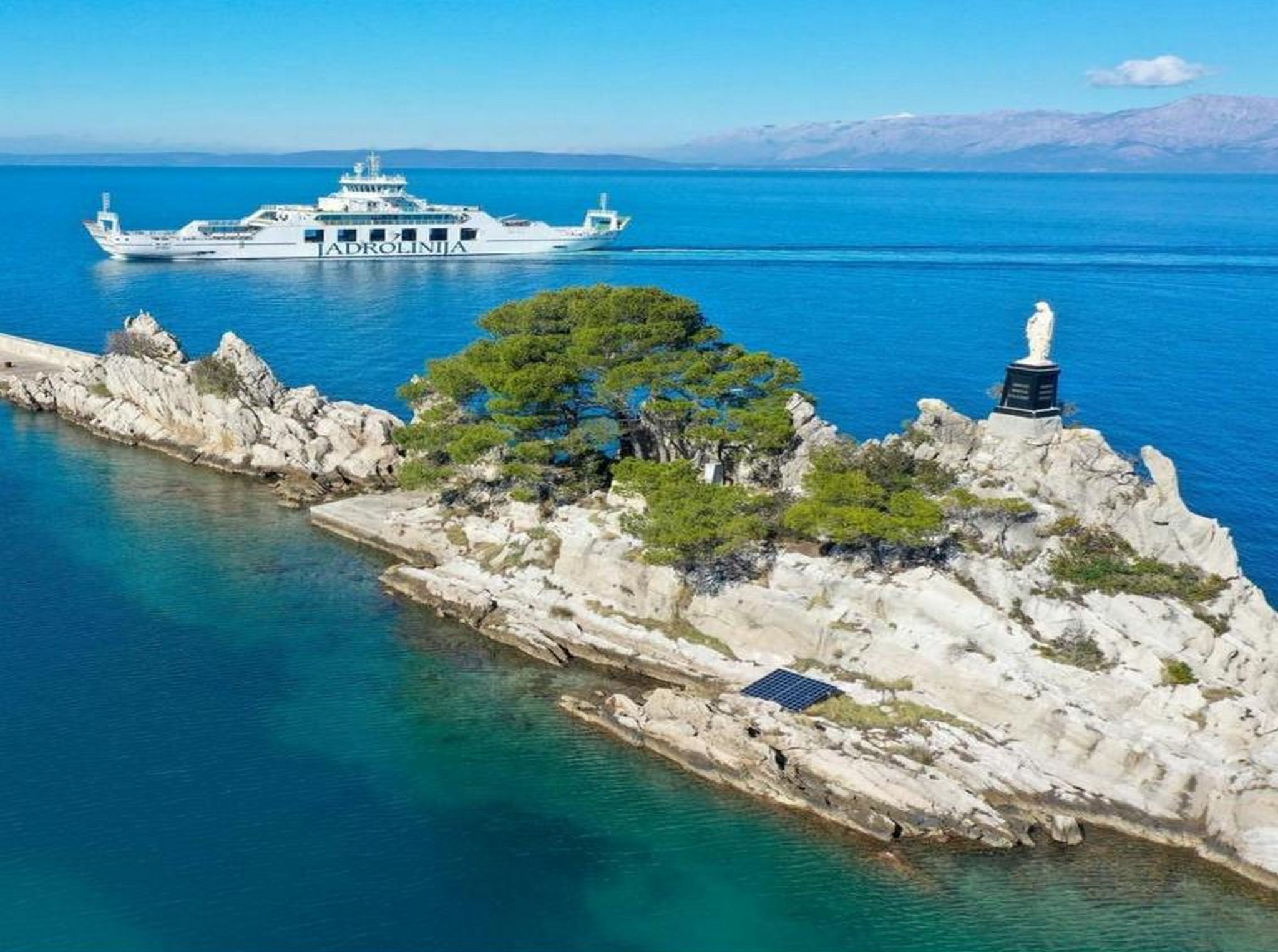 Un paysage côtier avec un terrain rocheux, une statue et, au loin, un bateau nommé « ADROLINJA ».