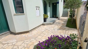 Un bâtiment dont l’entrée est pavée de pierres, avec une porte verte et un petit jardin agrémenté de fleurs violettes et rouges.
