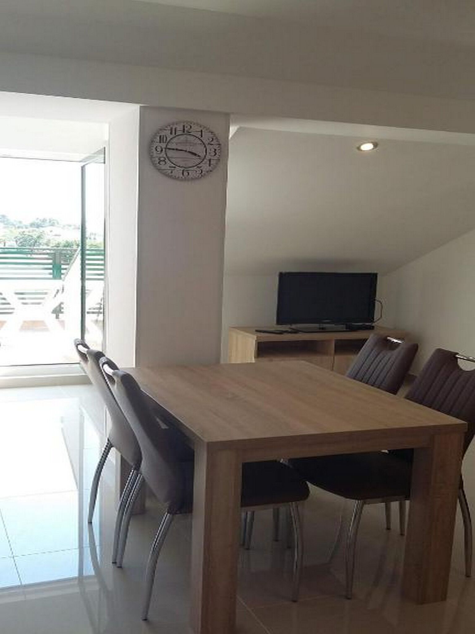 Un comedor con una mesa y sillas de madera, un televisor sobre un mueble bajo y un reloj en la pared.