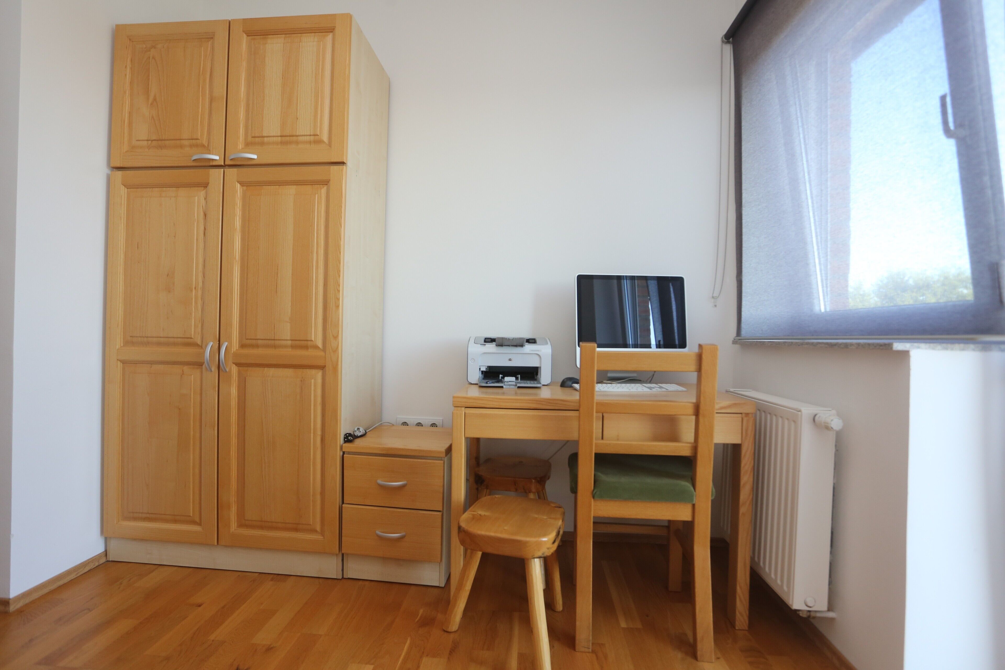 A room with a wooden wardrobe, a desk with a printer, a chair, and a radiator.