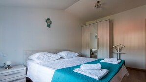 Una cama bien hecha con ropa blanca y una manta turquesa, una mesita de noche blanca con una lámpara, y un espejo grande en la pared.