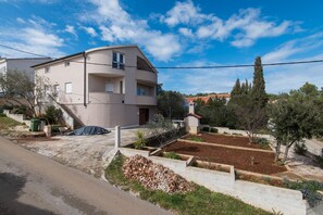 Un immeuble résidentiel à plusieurs étages, doté d’un balcon, d’une allée et d’un jardin agrémenté d’arbres et d’arbustes.