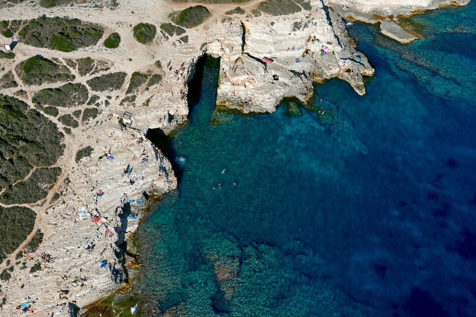 An aerial view of a rocky coastline with clear blue waters, featuring a rugged terrain with scattered vegetation and a few people enjoying the beach.