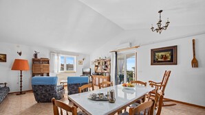 Un coin repas avec une table dressée pour quatre personnes, entourée de chaises en bois. On aperçoit une bibliothèque remplie de divers objets et, au fond, un téléviseur.