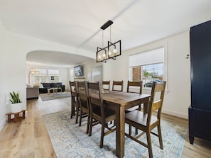 A dining area with a wooden table and chairs, a hanging light fixture, and a view of the kitchen in the background.