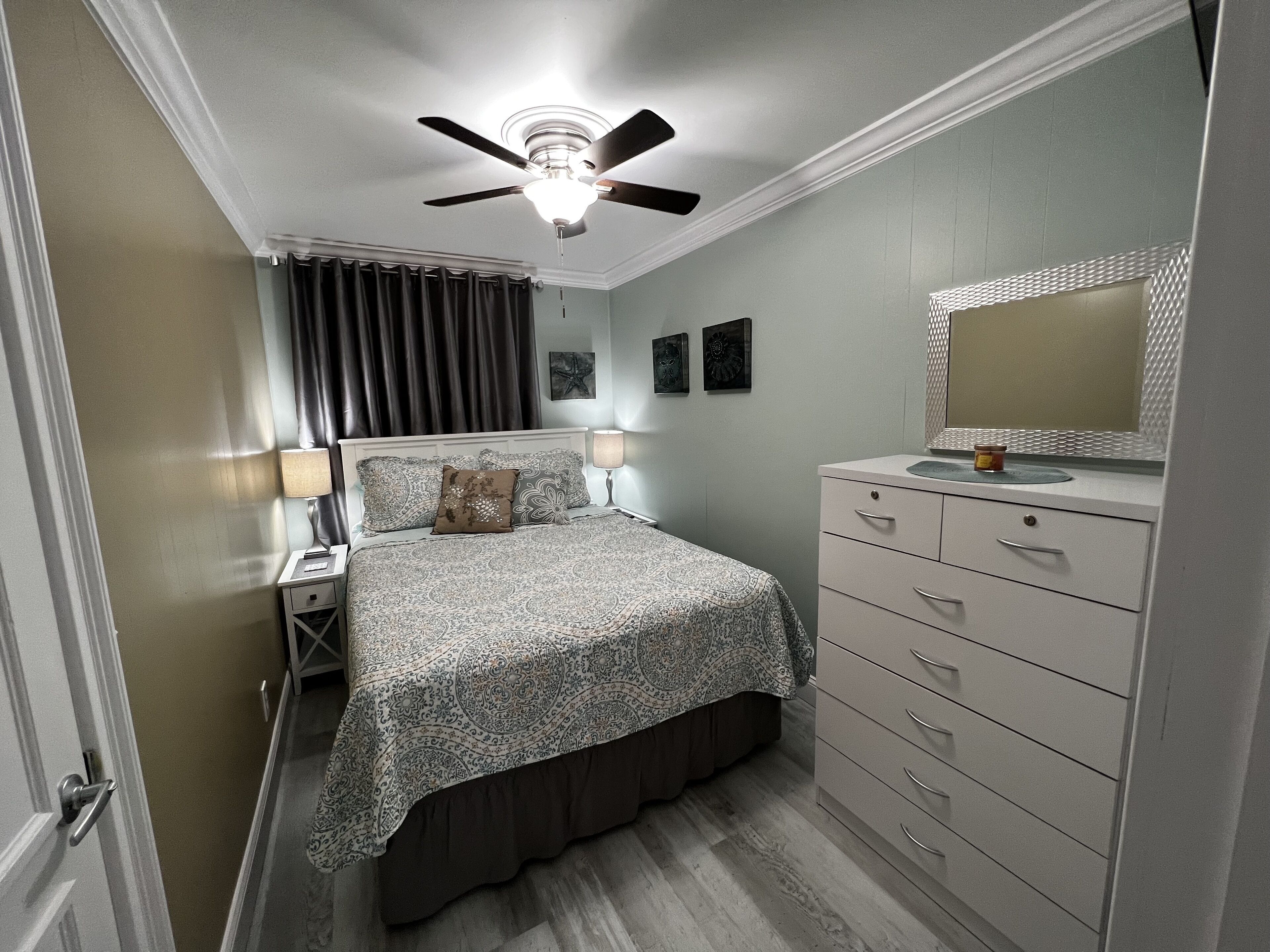 A bedroom with a bed, bedside lamps, a nightstand, a dresser, a ceiling fan, and a mirror.