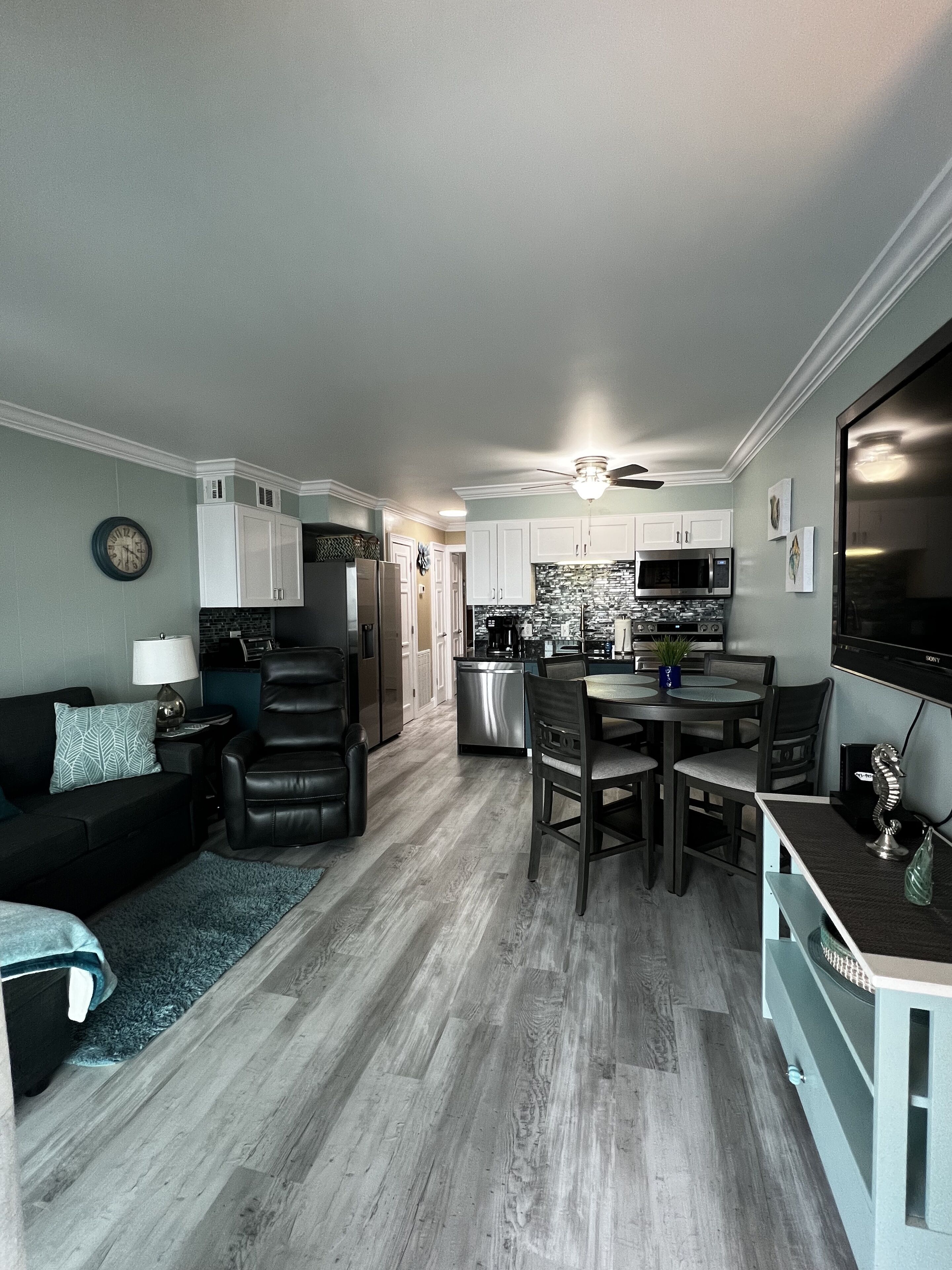 A modern kitchen with white cabinets, a dining area with a table and chairs, a black leather sofa, and a flat-screen TV.