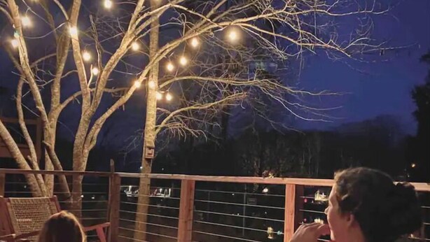 Une terrasse agrémentée d’un foyer et de guirlandes lumineuses, entourée d’arbres.