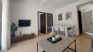 A modern living room with a flat-screen TV, a coffee table with a plant and candle, a dining area with chairs, and a hallway leading to a door.