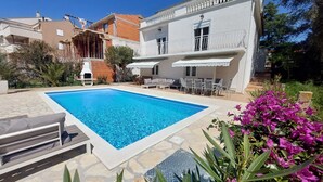 A swimming pool surrounded by stone tiles, with outdoor seating and a white building in the background.