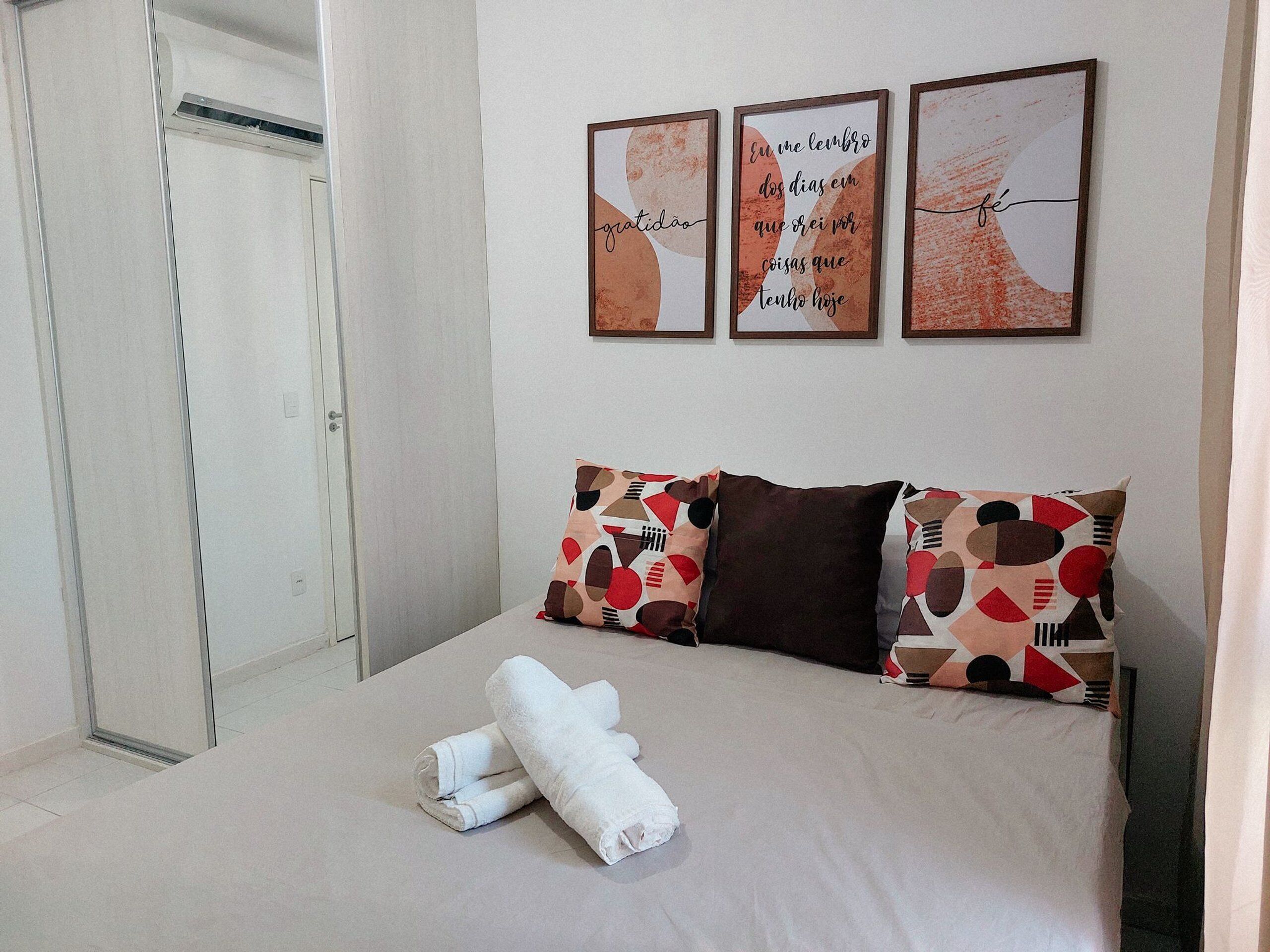 A neatly made bed with a beige bedspread, two pillows with a geometric pattern, and two rolled white towels. There are three framed pictures on the wall above the bed.