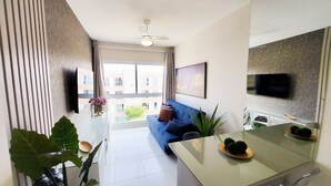 A modern living room with a blue sofa, a dining table with two chairs, a ceiling fan, and a TV mounted on the wall. - Flats no Muro Alto Clube (Ipojuca)