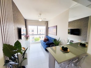 A modern living room with a blue sofa, a dining table with two chairs, a ceiling fan, and a TV mounted on the wall. - Flats no Muro Alto Clube (Ipojuca)