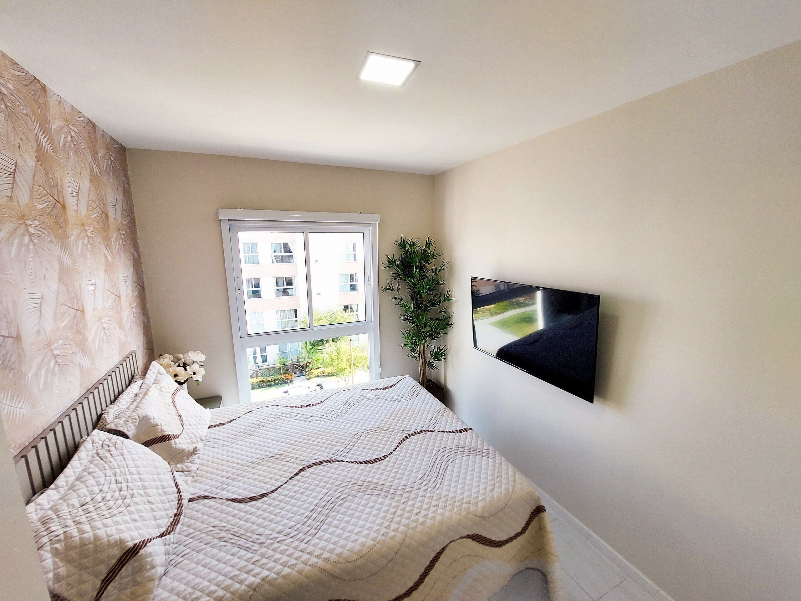 Un dormitorio con una cama, una ventana, una planta y un televisor montado en la pared.
