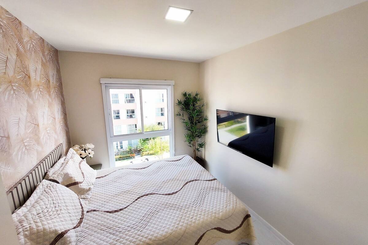A bedroom with a bed, a window, a plant, and a TV mounted on the wall.