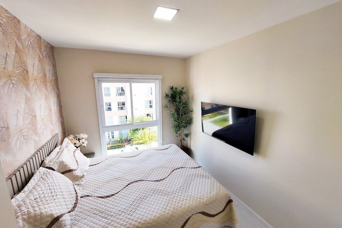 Un dormitorio con una cama, una ventana, una planta y un televisor montado en la pared.