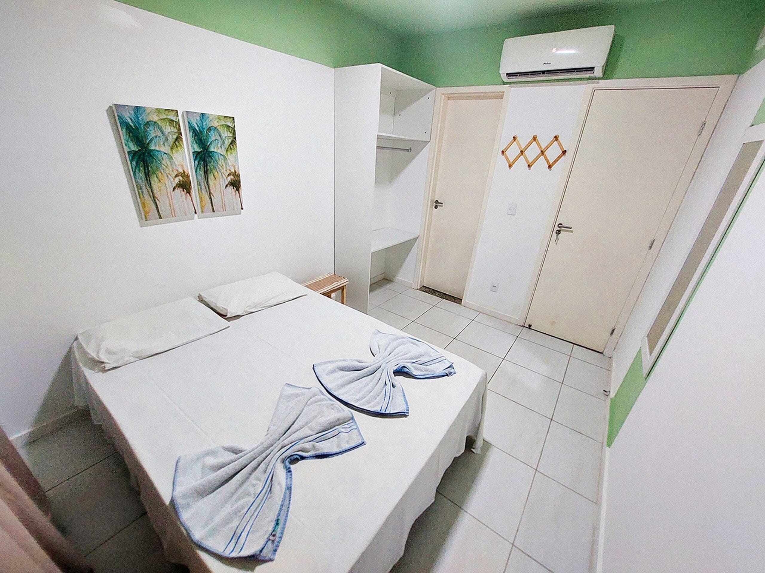 A hotel room with a bed, a small table, a chair, a wardrobe, and two wall-mounted paintings.