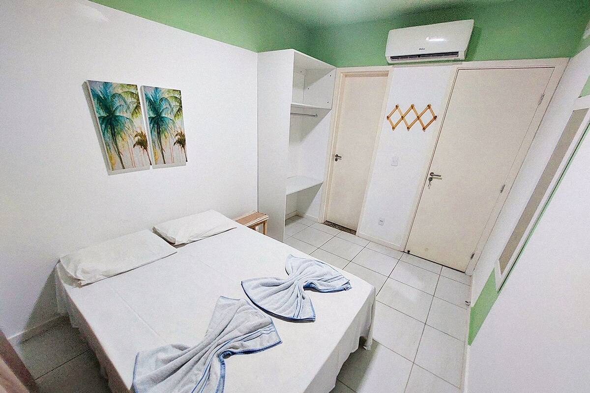 A hotel room with a bed, a small table, a chair, a wardrobe, and two wall-mounted paintings.