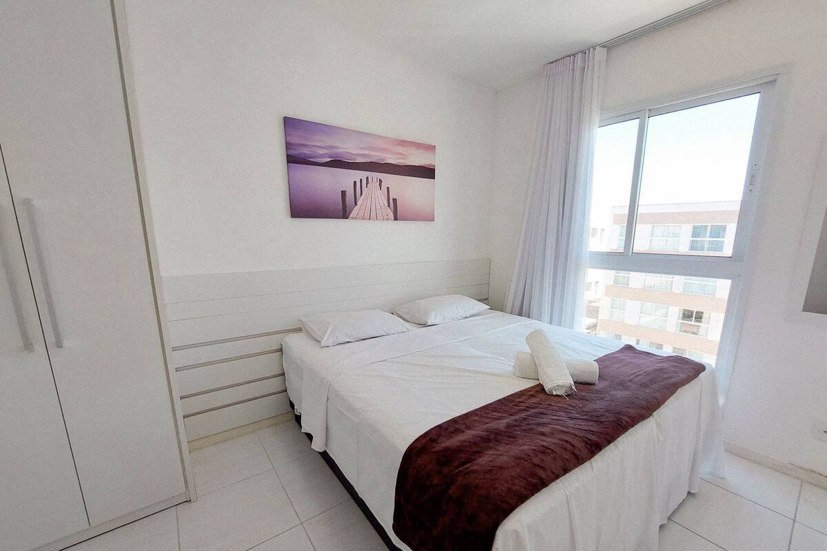 A hotel room with a bed, a window with curtains, a painting on the wall, and a view of buildings outside.
