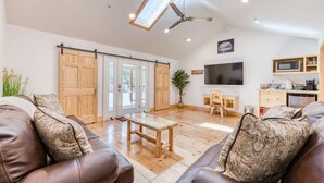 A spacious living room with a wooden floor, leather sofas, a wooden coffee table, a ceiling fan, a TV, and a kitchen area with a microwave and cabinets. - Cottage Like Suite w/ Private Hot Tub (Bass Lake)