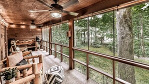 A wooden cabin with a balcony, furnished with a sofa and chairs, and large windows offering a view of the forest. - Magical Forest Lodge-HotTub-Pool Table-Fireplace-Creek (Ellijay)