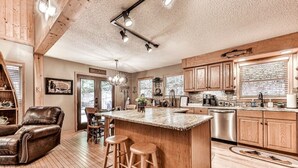A spacious kitchen with wooden cabinets, a large island with stools, and a dining area with a leather chair. - Magical Forest Lodge-HotTub-Pool Table-Fireplace-Creek (Ellijay)