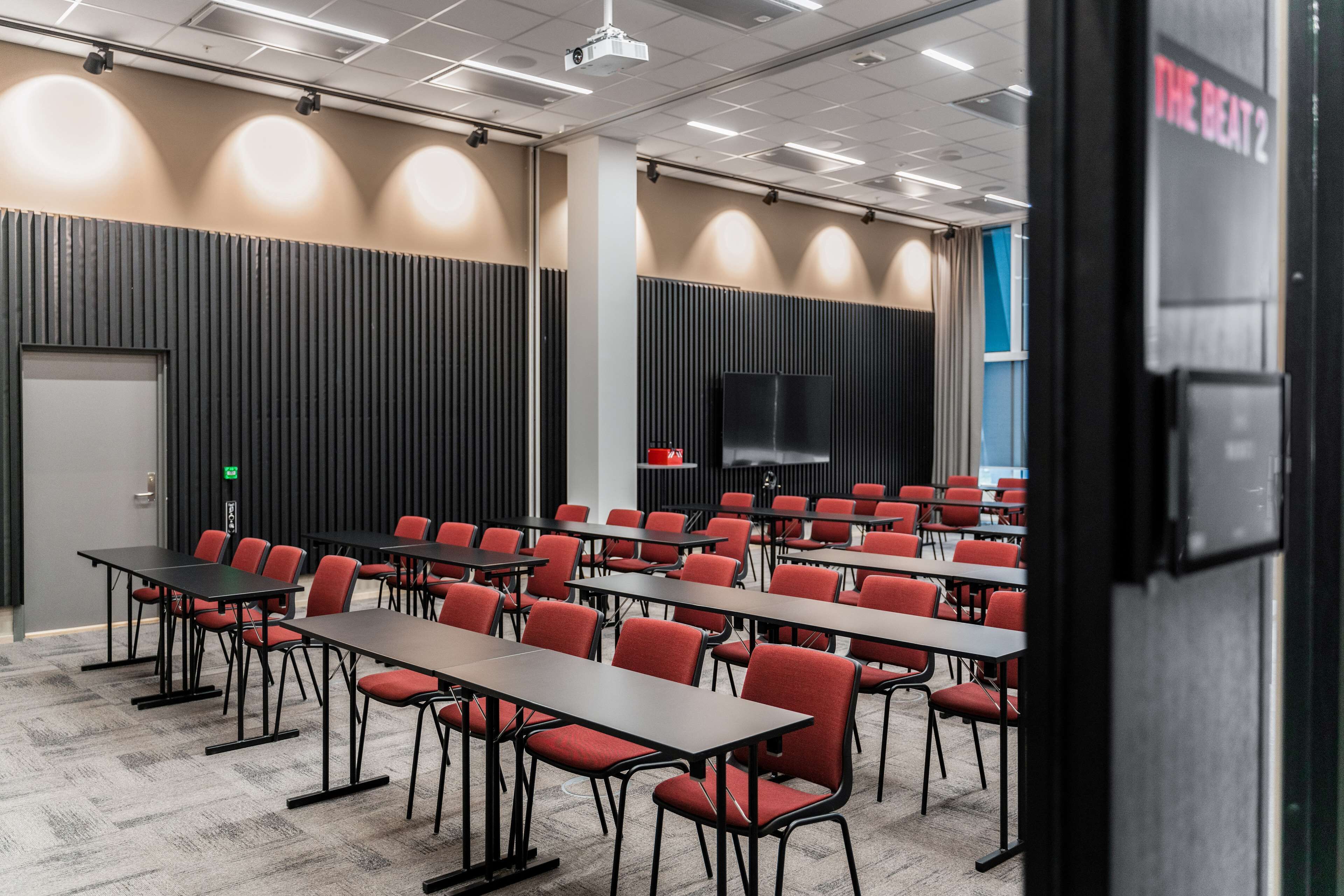 A conference room with rows of tables and chairs, a large screen, and a door with a sign.