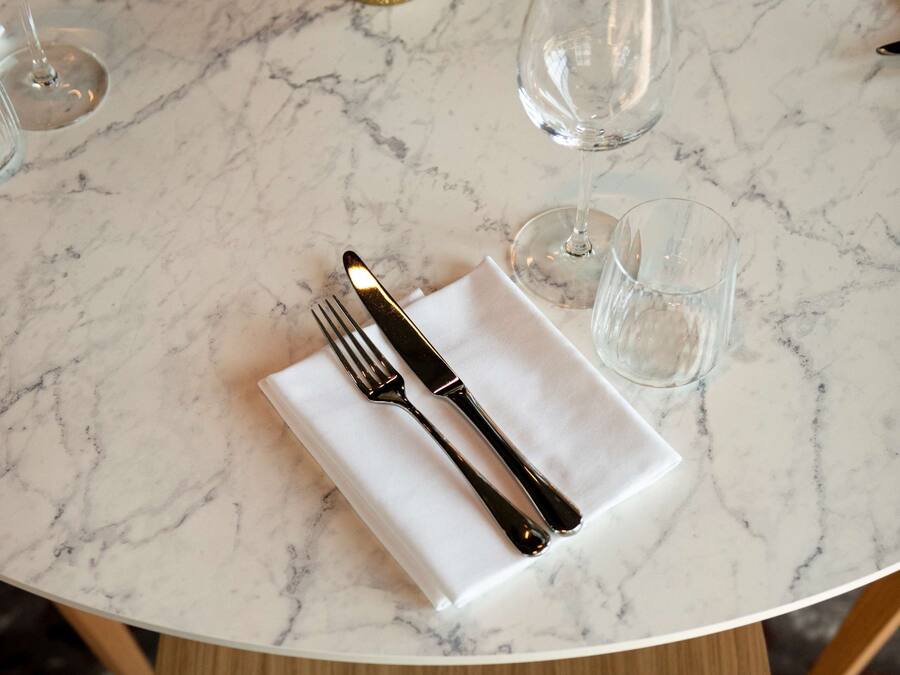 Una mesa de mármol con un mantel blanco, un tenedor y un cuchillo. Hay dos vasos y un salero sobre la mesa.