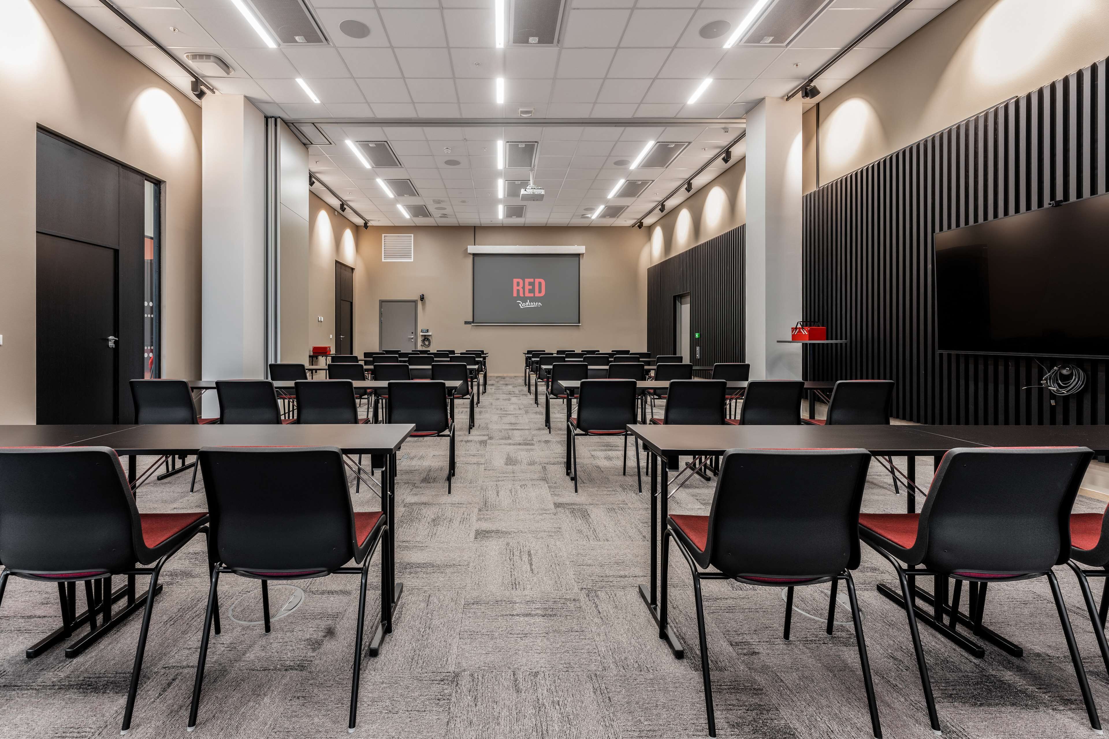 A conference room with rows of tables and chairs, a projector screen, and a large flat-screen TV.