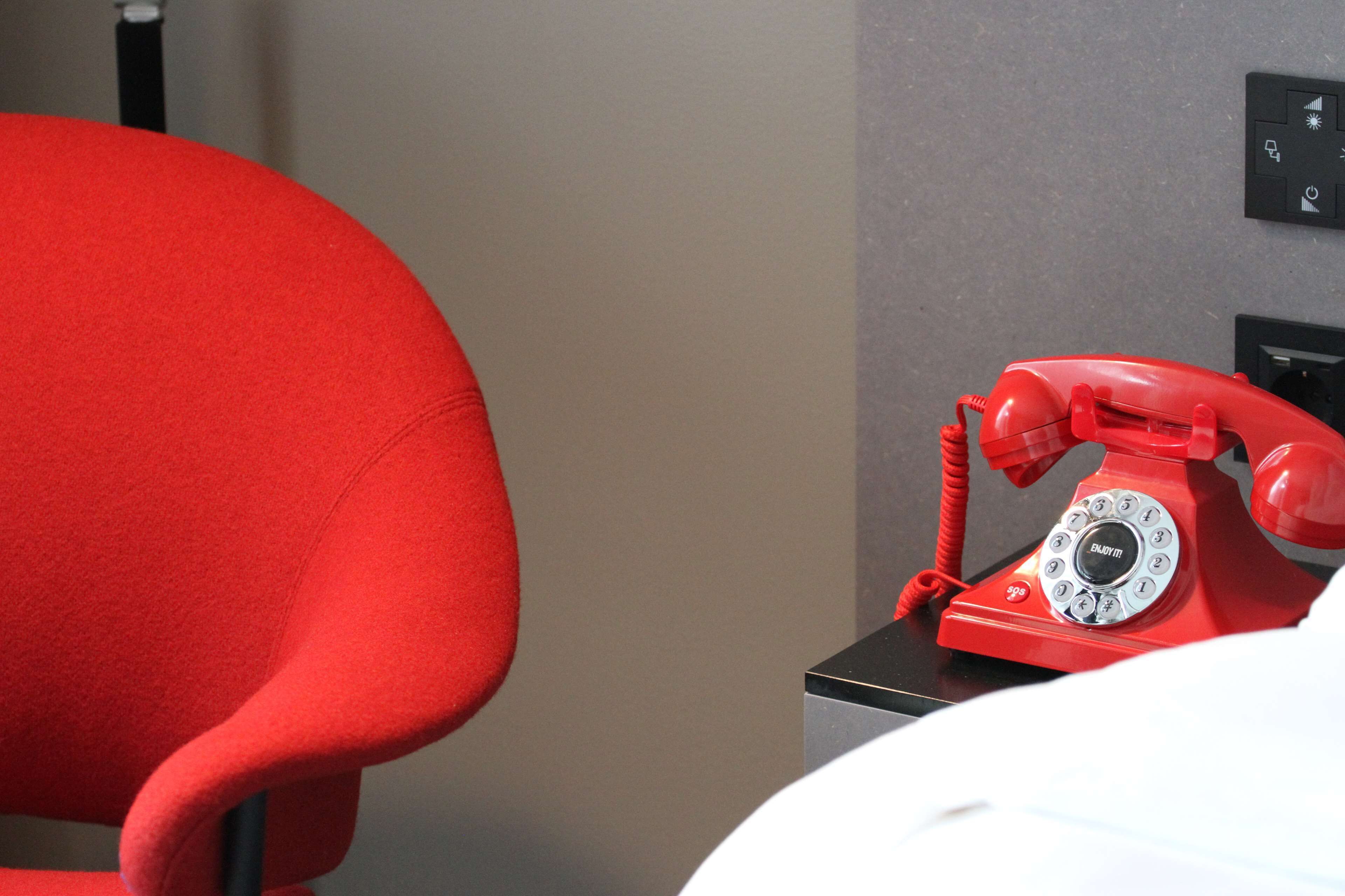 A red telephone on a black base, a red chair, and a white bed in a room.