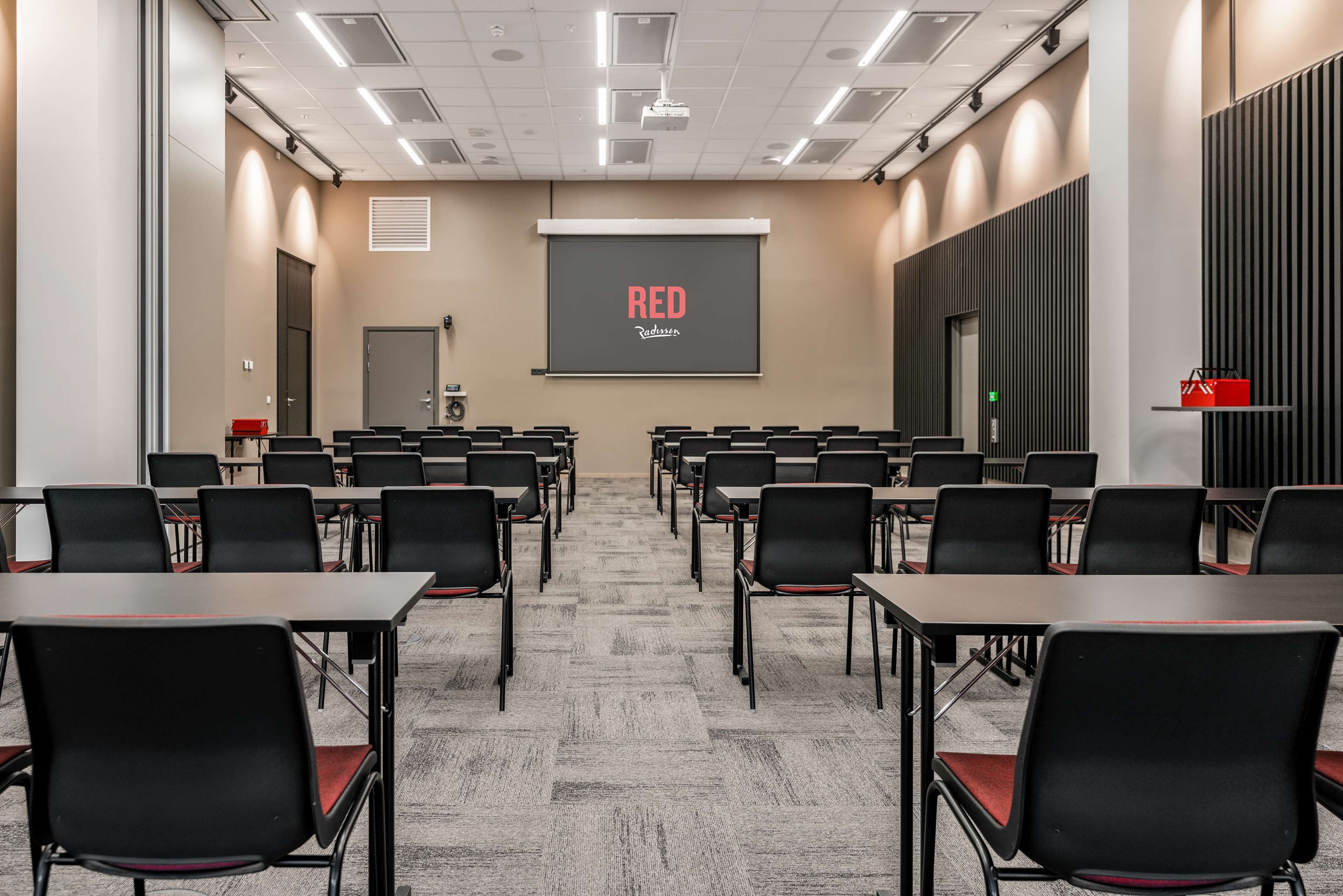 A conference room with rows of tables and chairs, a projector screen with the text "RED Radisson", and a door at the far end.