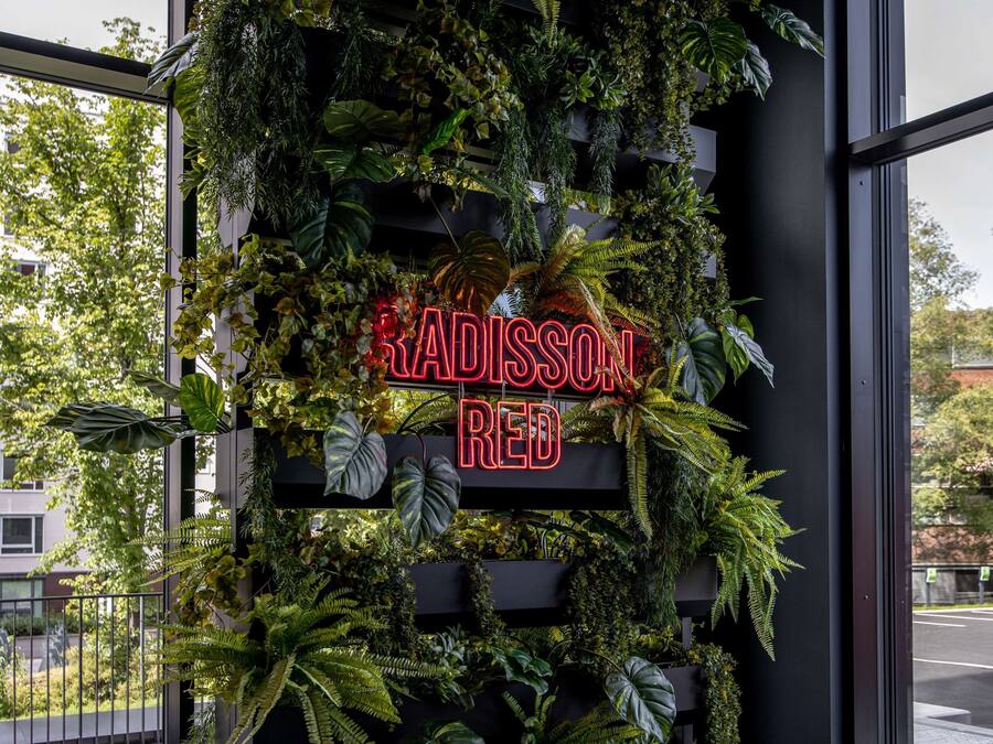 Seinä, jossa on neonvaloin valaistu kyltti, jossa lukee ”RADISSON RED”, ja sen ympärillä on vehreää kasvillisuutta.