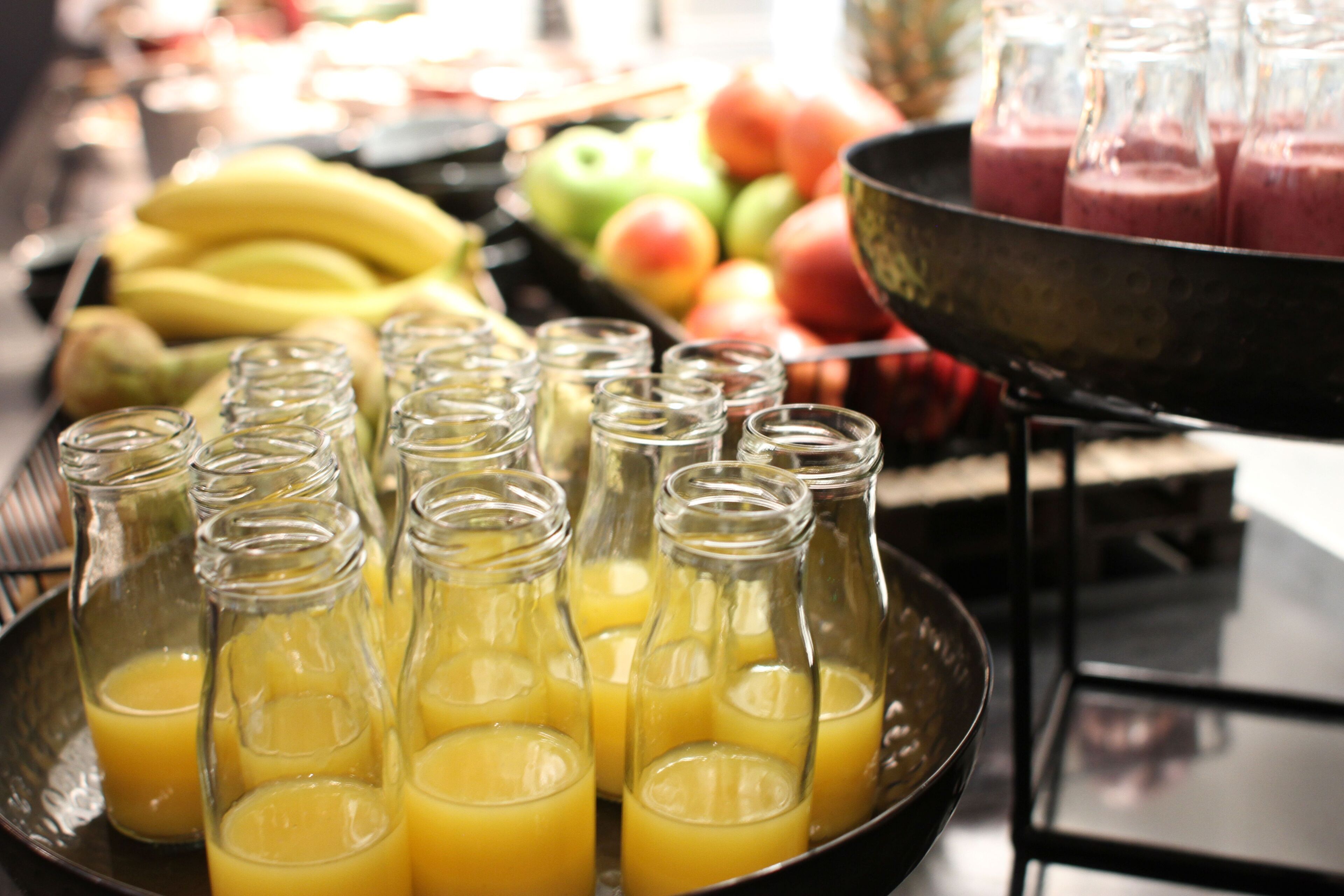 A tray of glass bottles filled with orange juice, a bowl of bananas, and a variety of fruits including apples and peaches.