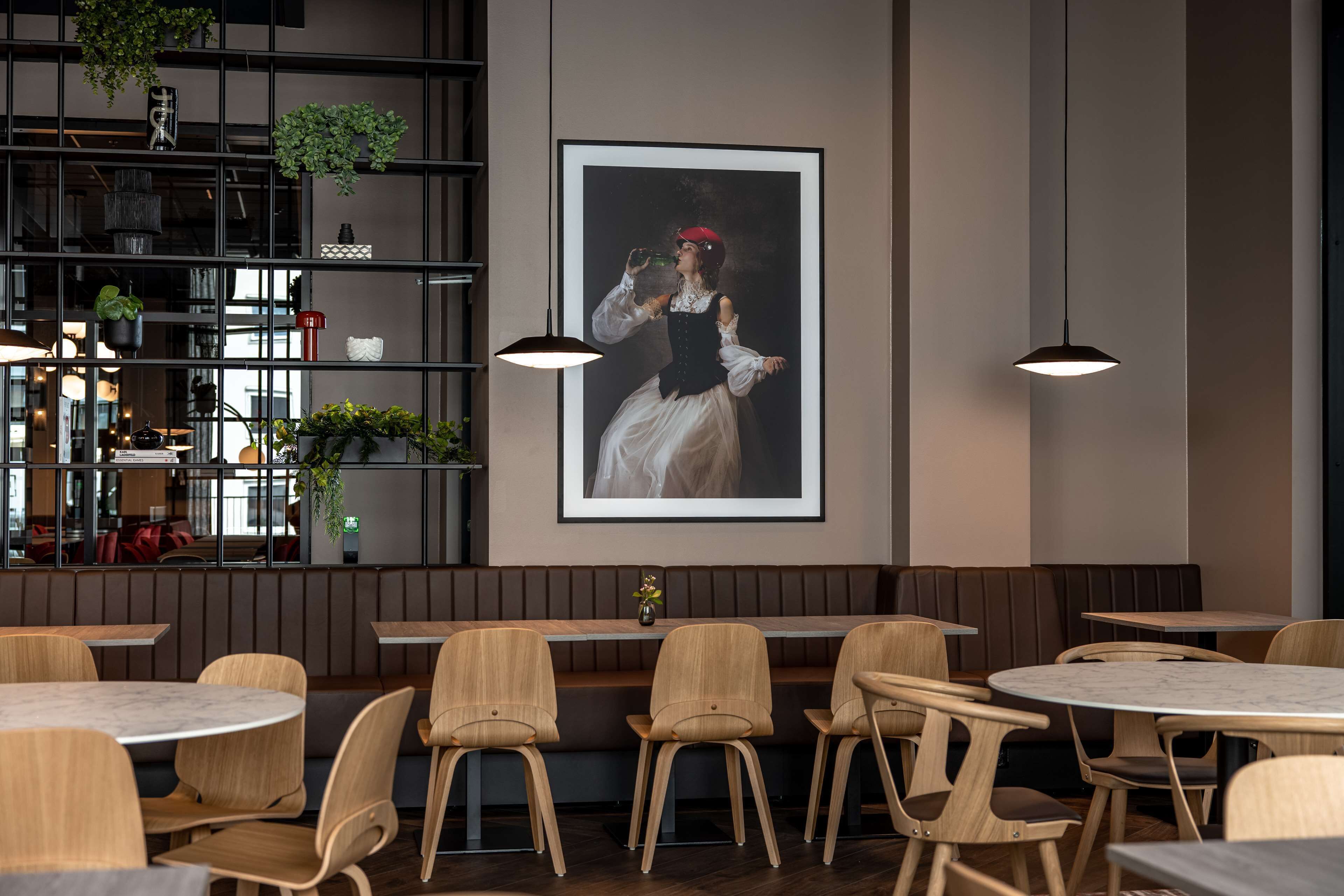 A restaurant interior with wooden tables and chairs, a large framed portrait on the wall, and hanging plants.