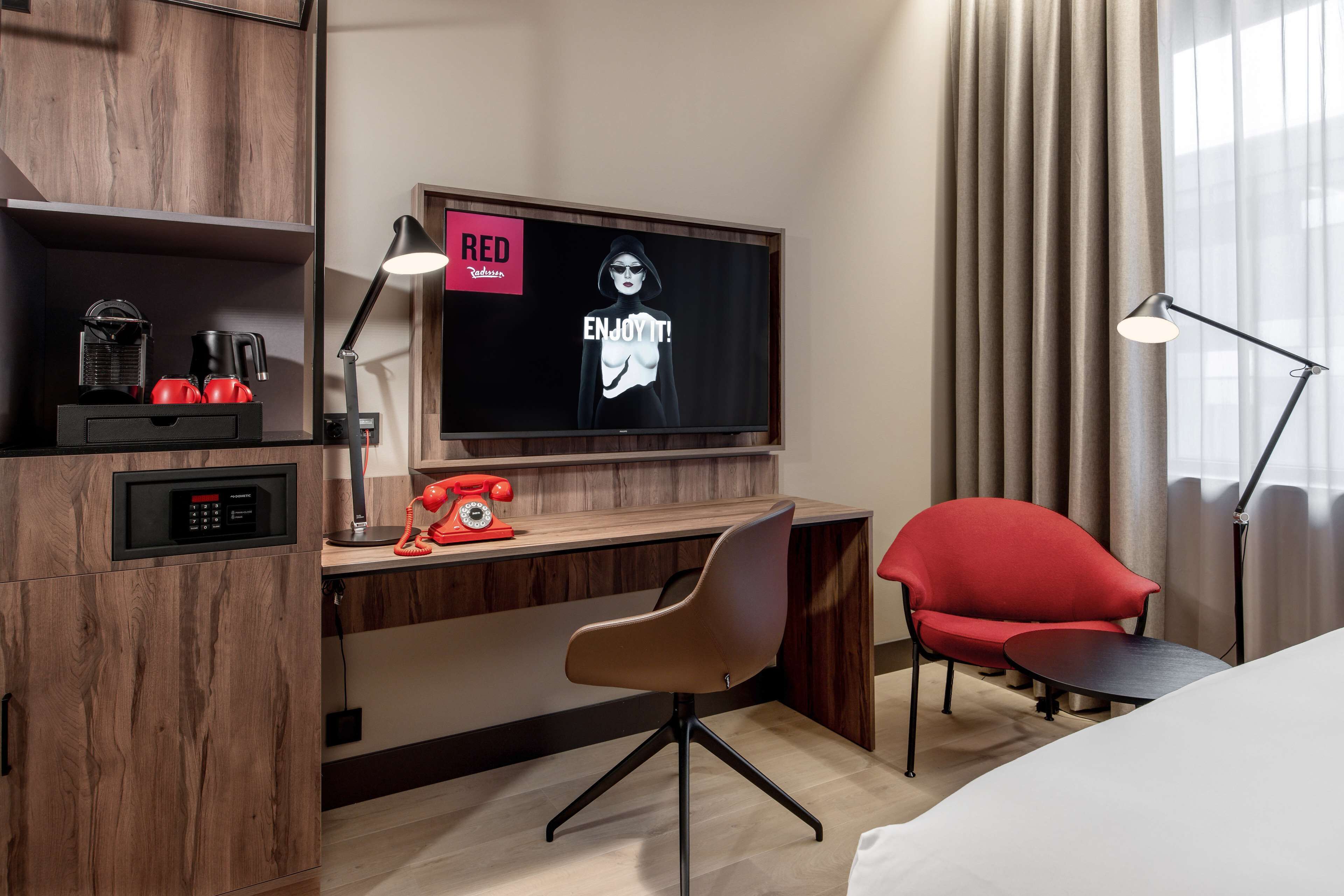 Una habitación de hotel con un escritorio de madera, un teléfono rojo, una silla moderna, un televisor y una lámpara.