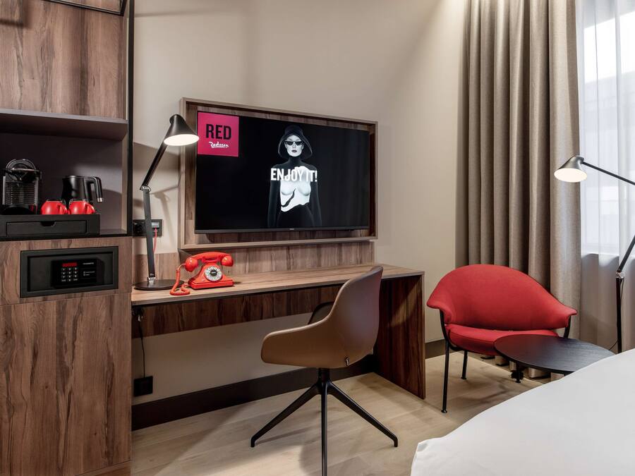 A hotel room with a wooden desk, a red telephone, a modern chair, a television, and a lamp.