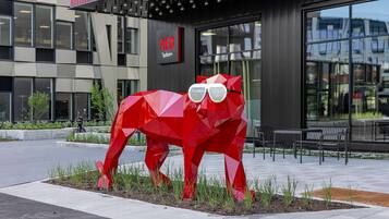 Une sculpture géométrique rouge représentant un chat portant des lunettes de soleil, placée devant un bâtiment sur lequel on peut lire l’inscription « RED ».