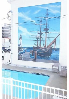 Um mural retrata um navio a vela em um cais, com um farol, uma piscina e um edifício branco com varandas.