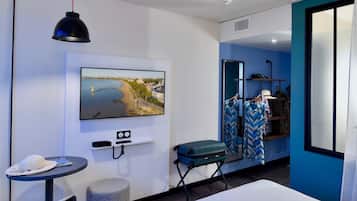 Habitación de hotel con cama, televisor montado en la pared, una mesita con un sombrero y una revista, maleta y espejo con ropa.