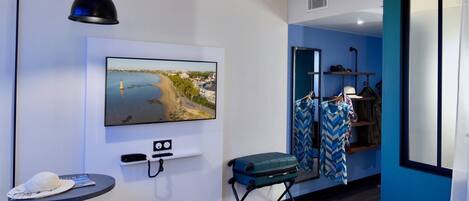Habitación de hotel con cama, televisor montado en la pared, una mesita con un sombrero y una revista, maleta y espejo con ropa.