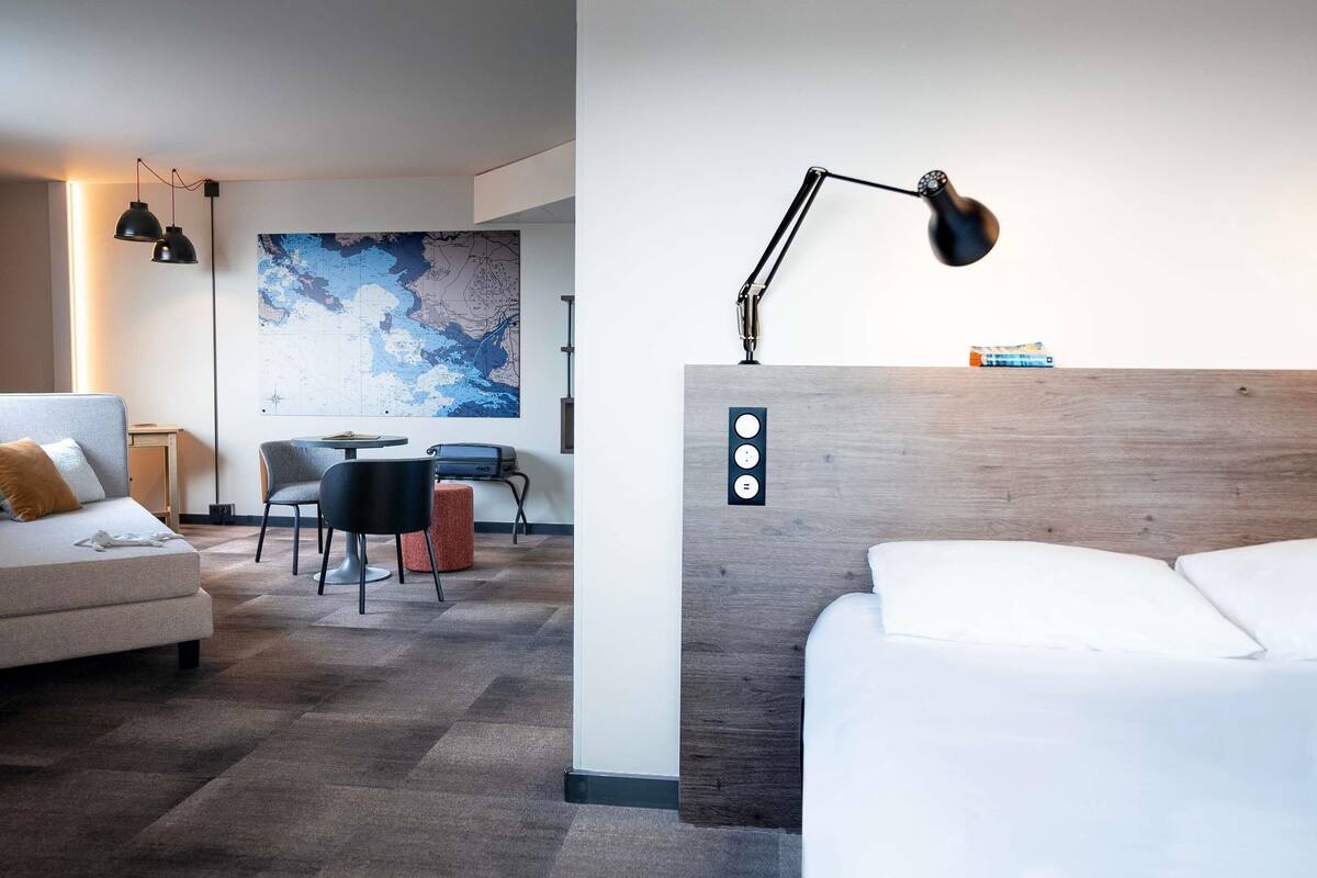 A hotel room with a bed, a desk, a chair, a map on the wall, and a lamp.