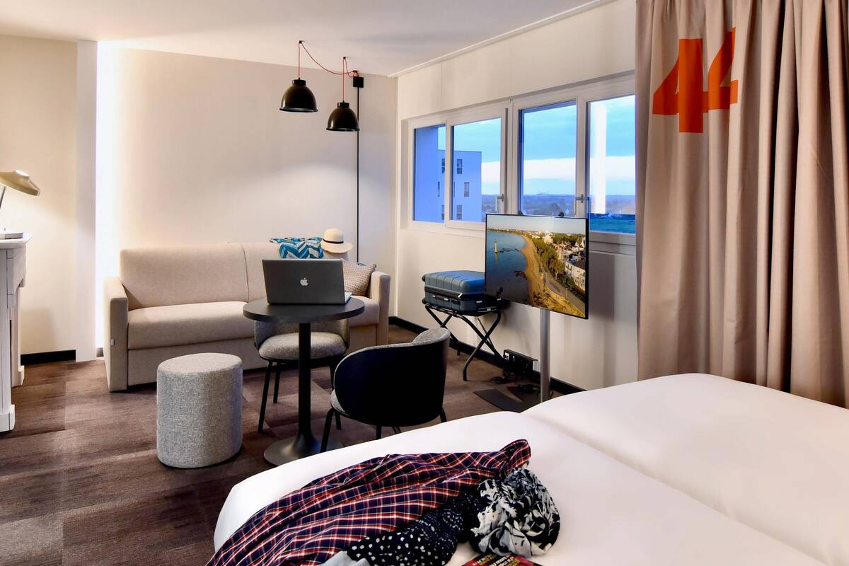 A hotel room with a bed, a desk with a laptop, a chair, a sofa, a TV, and a window with curtains.