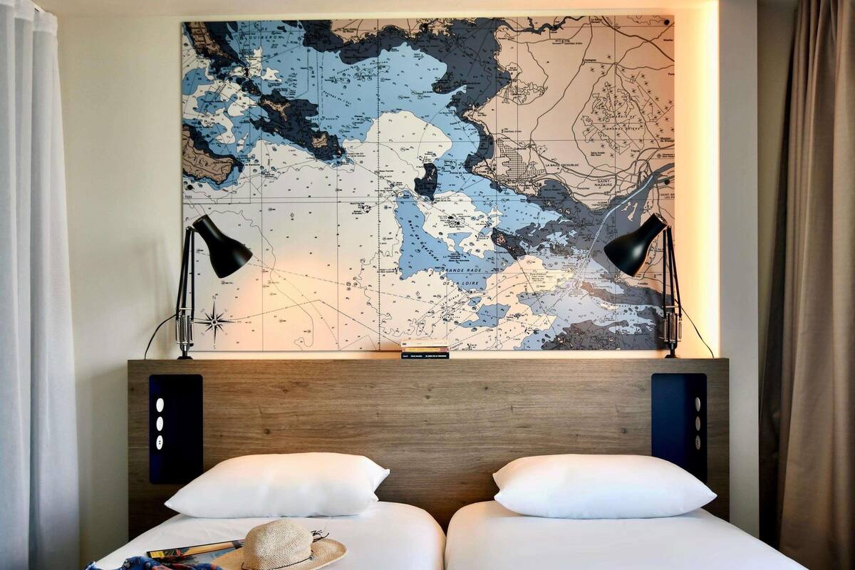 A hotel room with a large map on the wall, two bedside lamps, a bed with white bedding, and a hat on the bed.