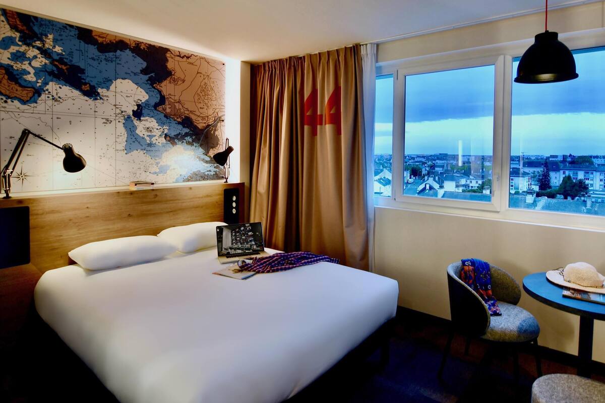 A hotel room with a large bed, a map on the wall, a desk with a laptop, a view of the city through the window, and a table with a hat.