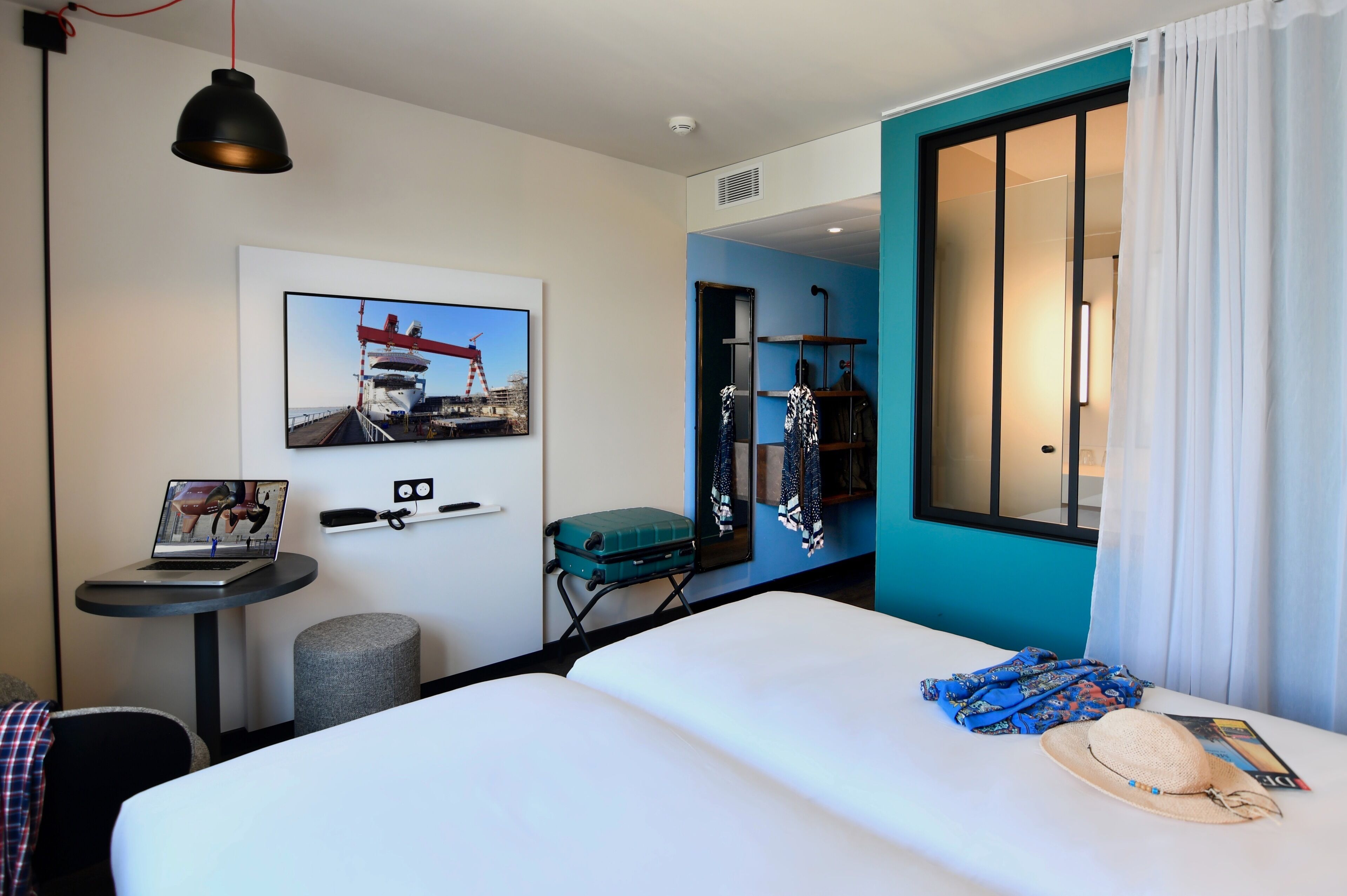 Una habitación de hotel con cama, escritorio, computadora portátil, maleta, sombrero y ventana con cortinas.
