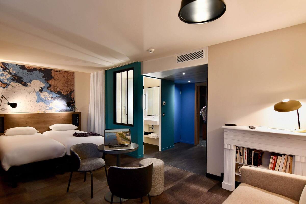 A hotel room with a bed, a desk with a laptop, a chair, a bookshelf, a map on the wall, and a fireplace.